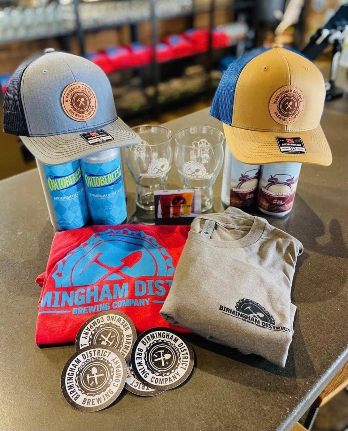Birmingham District Brewing full merch collection flat lay including embroidered leather patch trucker caps, screen printed t-shirts, beer cans, and pint glasses, custom produced by Presto Embroidery in Birmingham AL